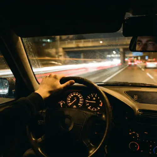Die lockere Hand eines übermüdeten Mannes am Lenkrad eines dunklen Wagens, umgeben von verschwommenen, grellen Autobahnlichtern.