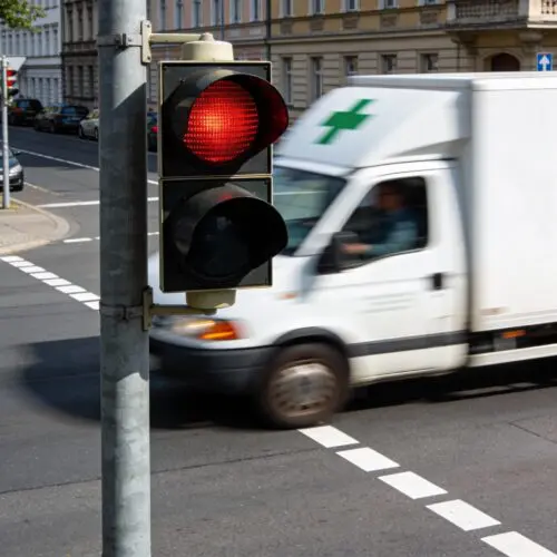 Ein weißes Apotheken-Lieferfahrzeug fährt eilig bei rot leuchtender Ampel über die Haltelinie einer Kreuzung.