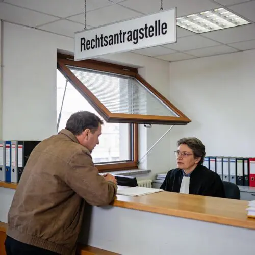 Ein laienhafter Mann spricht am Schalter der Rechtsantragstelle eines Gerichts mit einem Justizbeamten.