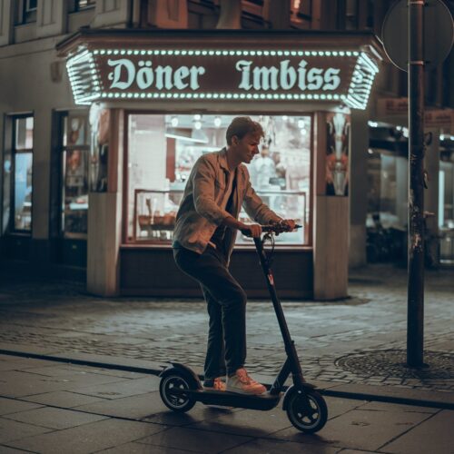 Student fährt unsicher auf E-Scooter vor beleuchtetem Dönerimbiss