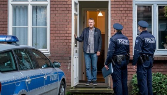 Mann in Bremer Reihenhaus steht verwirrt im Türrahmen, während zwei Polizisten davor stehen.