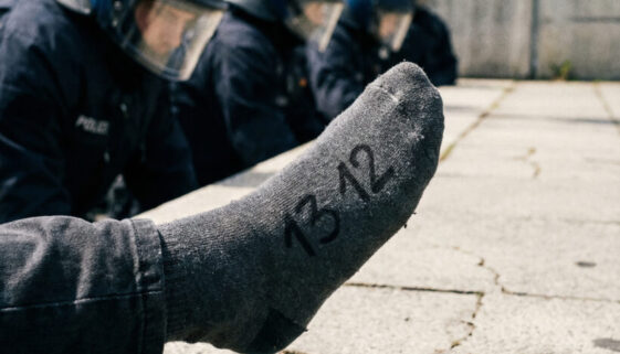 Nahaufnahme der Zahl „13 12“ auf der angehobenen Socke eines Demonstranten, konfrontativ auf uniformierte Polizeibeamte gerichtet.