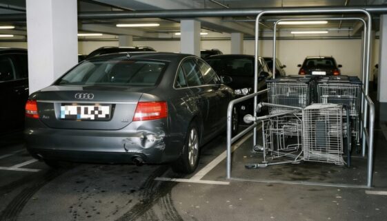 Ein moderner PKW mit Kollisionsschaden am Heck fährt eilig am verbogenen Einkaufswagen-Gestell vorbei.