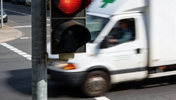 Ein weißes Apotheken-Lieferfahrzeug fährt eilig bei rot leuchtender Ampel über die Haltelinie einer Kreuzung.