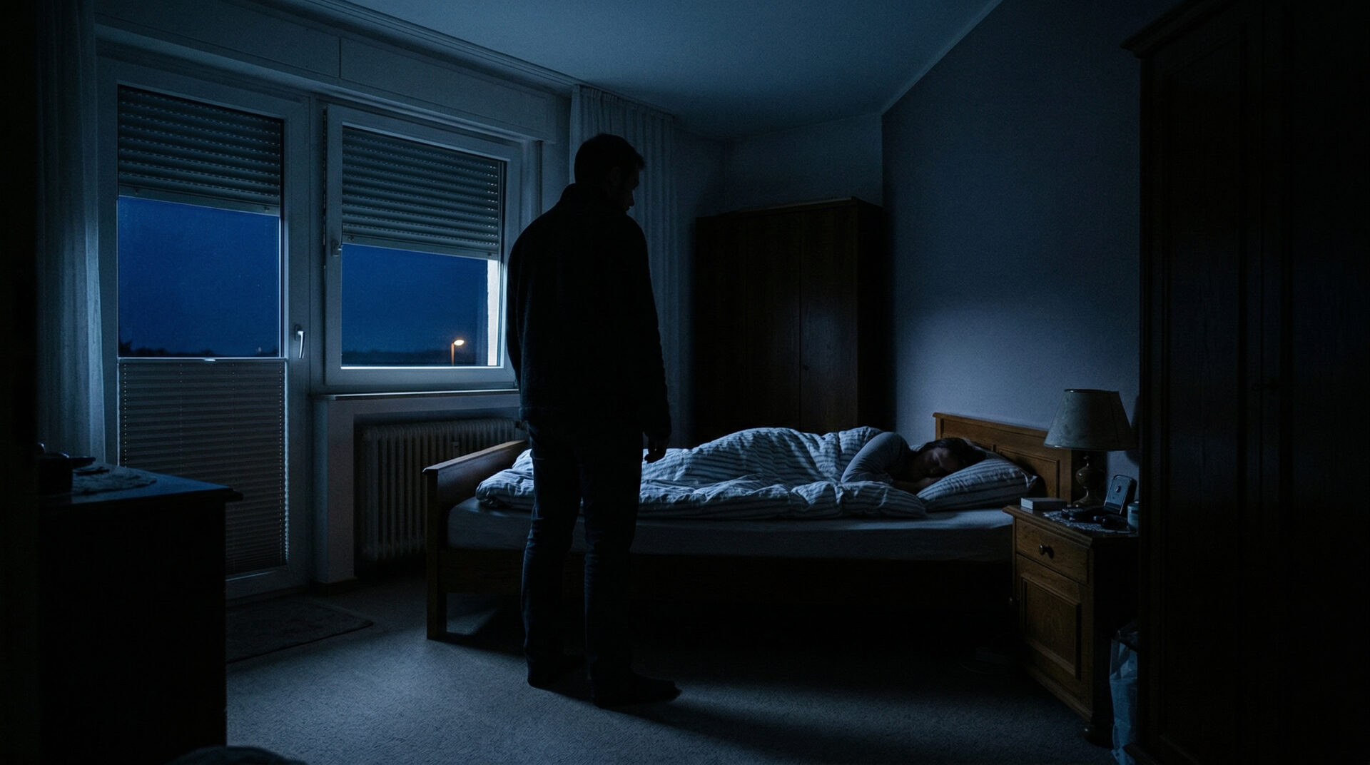 Männliche Silhouette steht im fahl beleuchteten Schlafzimmer neben einem Bett, in dem eine Person ruhig schläft.