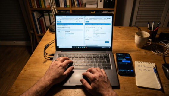 Hände an Laptop mit Online-Banking-Fenstern, Smartphone und Notizzettel mit Provisionsberechnung auf Schreibtisch.