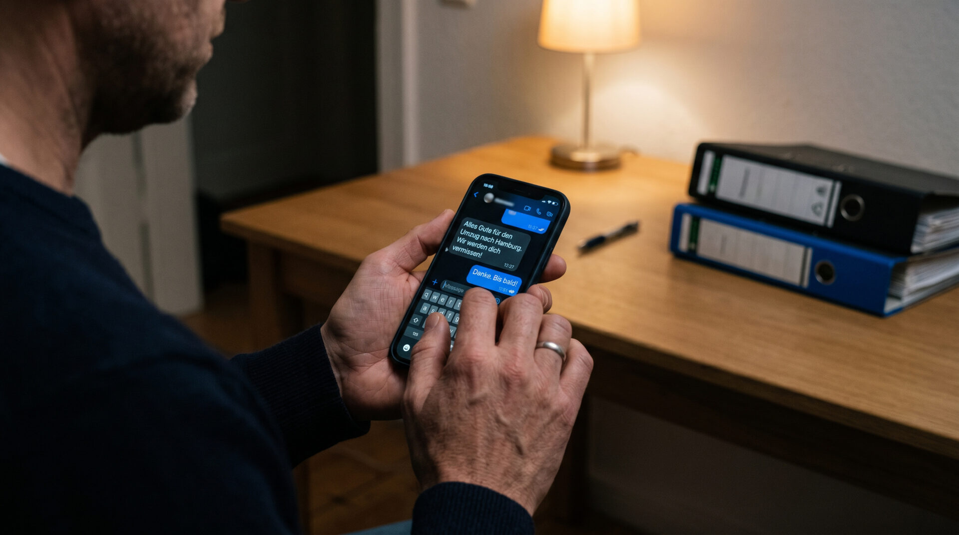 Hände halten ein Smartphone mit einer Chat-Nachricht in einem nüchternen Raum; im Hintergrund Aktenordner.