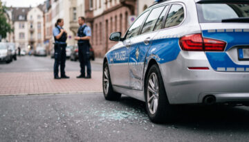 Defektes Polizeifahrzeug mit Dellen an der Seitenwand liegt auf einer normalen Stadtstraße, Glassplitter am Asphalt.